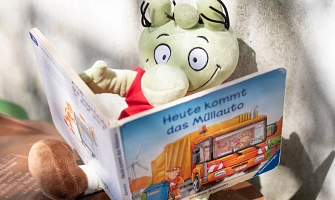 Eine Handpuppe von einem kleinen grünen Olchi sitzt in der Sonne und ließt ein Buch. Das Buch heißt "Heute kommt das Müllauto".